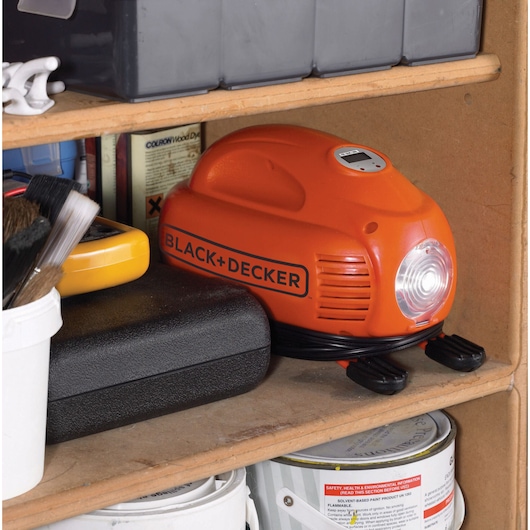 An orange BLACK+DECKER portable air compressor is placed on a wooden shelf next to other tools, brushes, and cans. The device features a built-in light and a digital display on the top.