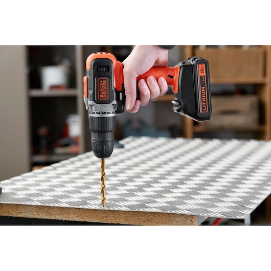 Photo of BLACKDECKER BCD003C1 cordless drill in use, drilling metal sheet, orange and black design, side angle.