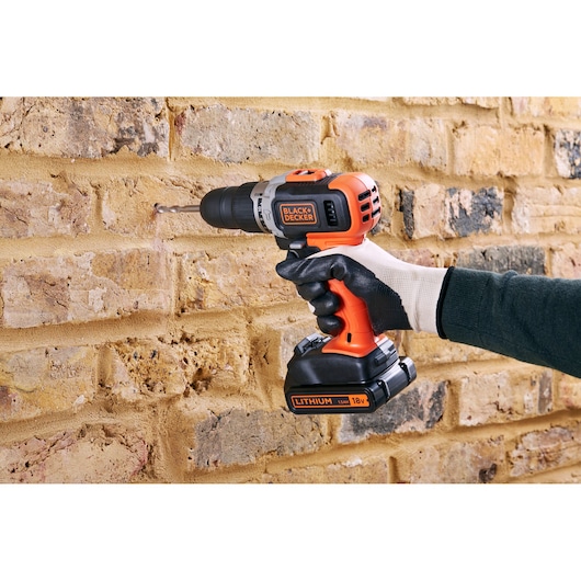 BLACK+DECKER BCD003C1 cordless drill with 18V lithium battery drilling into a brick wall, gloved hand holding.