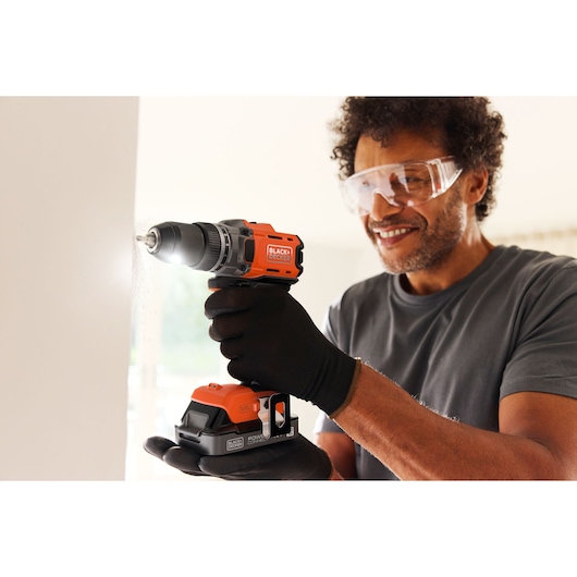 Person uses a BLACK+DECKER cordless drill to drill into a wall, wearing gloves and safety goggles.