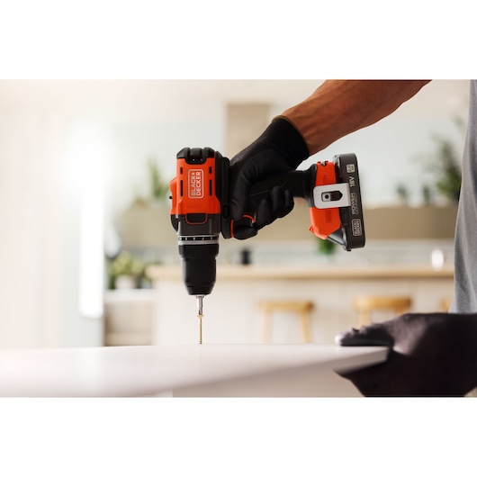 BLACK+DECKER cordless drill in use, drilling into a white surface indoors with a gloved hand.