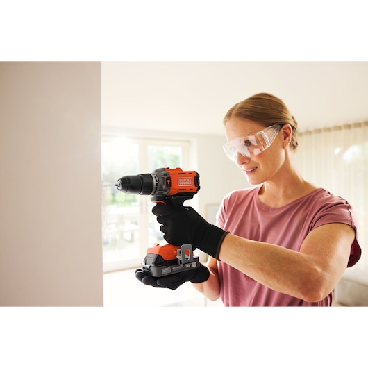 Person using a BLACK+DECKER cordless drill with safety glasses and gloves to drill into an indoor wall.