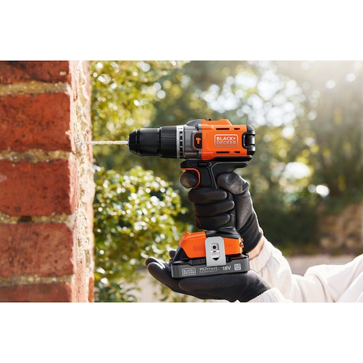 A BLACK+DECKER cordless drill in use, drilling into a brick wall outdoors, held by a gloved hand.