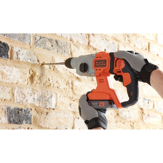 BLACKDECKER BCD900B cordless hammer drill held by gloved hands drilling into a brick wall, side angle.