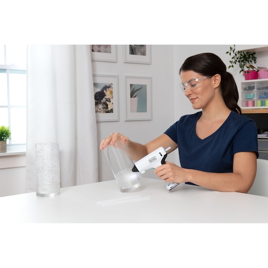 BLACKDECKER BCGL115 glue gun held by person applying glue to clear glass vase in craft workspace.