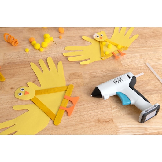 BLACK+DECKER BCGL115 cordless hot glue gun on a table with yellow paper bird crafts and pom-poms, top-down view.