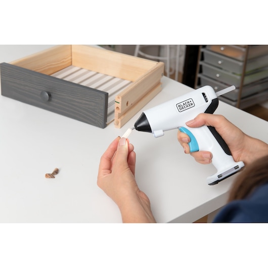 BLACKDECKER BCGL115 cordless hot glue gun held near small wooden dowel, drawer in background.