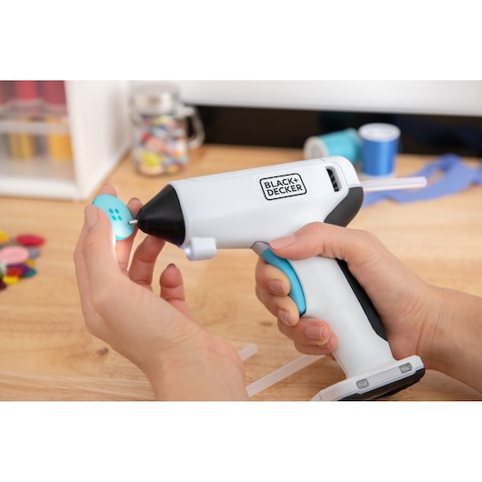 BLACK+DECKER BCGL115 glue gun held, applying glue to blue button in crafting setup.