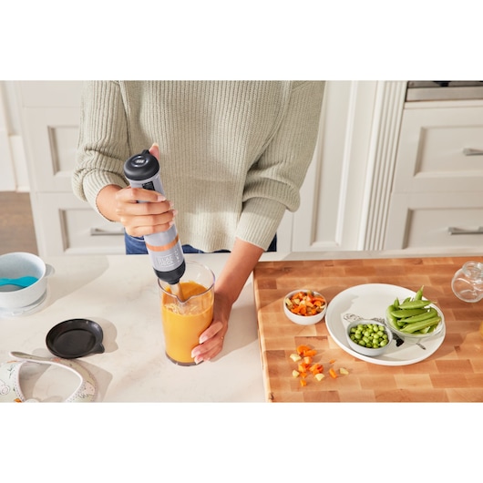 Photo of person using BLACK+DECKER BCKM1011KGF hand blender to mix orange liquid in glass on kitchen counter.
