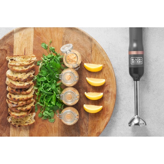 BLACK+DECKER BCKM1016KSG hand blender beside a wooden board with grilled bread, greens, sauce jars, and lemon wedges, top view.