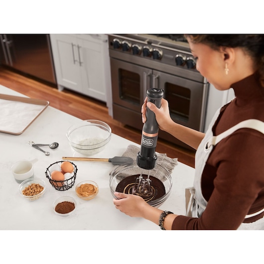 Photo of a person mixing batter in a glass bowl with a BLACK+DECKER BCKM101HM hand mixer in a kitchen.