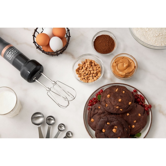 BLACK+DECKER BCKM101HM hand mixer with beaters on a white surface, surrounded by baking ingredients and chocolate cookies.