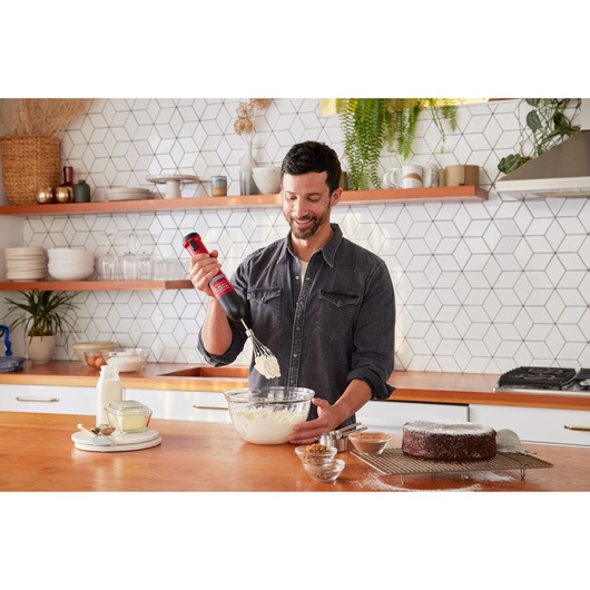 Photo of a person using a BLACKDECKER BCKM101WHFF hand mixer to whisk ingredients in a glass bowl on a kitchen counter.