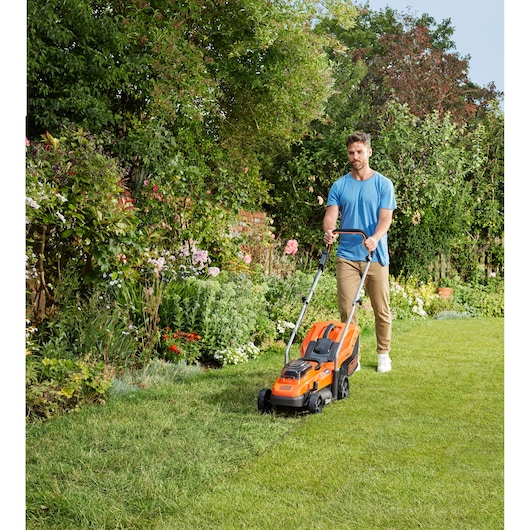 BLACKDECKER BCMW3318L2 lawn mower in use on grass, orange design, person pushing it in a garden setting.