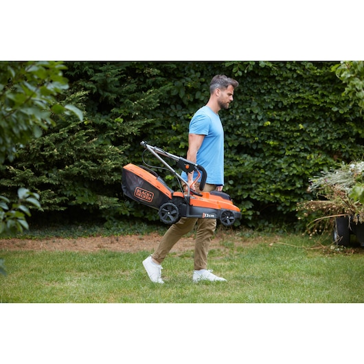 Photography of BLACKDECKER BCMW3318L2 lawn mower being carried outdoors by a person in casual wear.