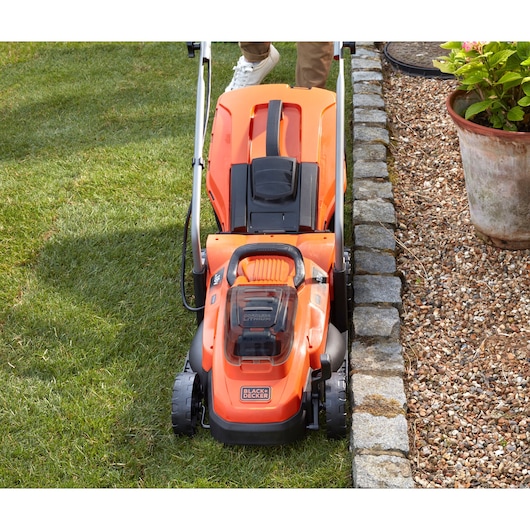 BLACKDECKER BCMW3318L2 lawn mower front angle, orange body, mowing grass near stone border.