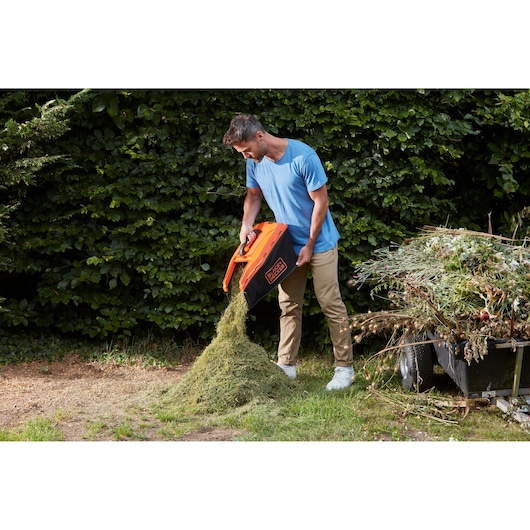 BLACKDECKER BCMW3318L2 lawn mower grass box being emptied outdoors onto a pile of grass clippings.