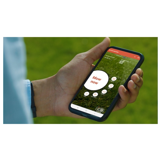 Photo of hand holding smartphone displaying BLACKDECKER BCRMW122 robotic mower app with 'Mow now' button.