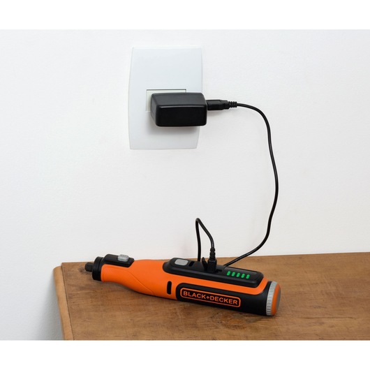 A BLACK+DECKER orange and black electric tool is shown charging on a wooden surface, connected to a power outlet through a black charger and cable.