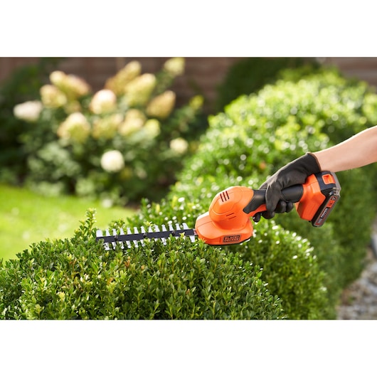 BLACKDECKER BCSS18B cordless shrub shear in use, orange and black, trimming green bushes outdoors.