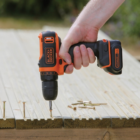 Photo of BLACK+DECKER BDCDD121 cordless drill being used to drive a screw into wooden boards outdoors.