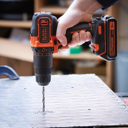 A hand holding a BLACK+DECKER 18V Lithium cordless drill, drilling a hole into a metallic surface.