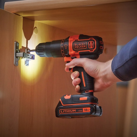 BLACK+DECKER BDCHD18S2A cordless drill being used to screw a cabinet hinge, 18V lithium battery visible.
