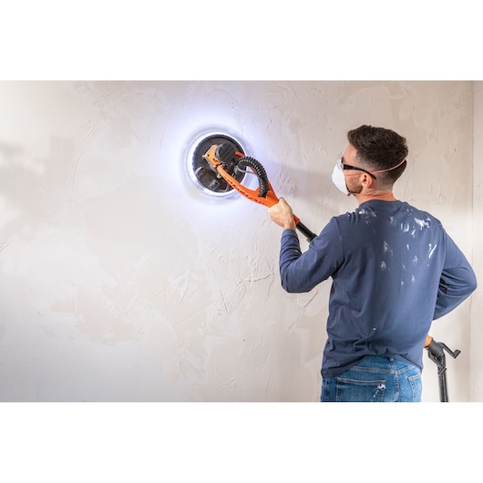 Person sanding a wall with a BLACK+DECKER LED drywall sander, wearing protective mask and goggles.