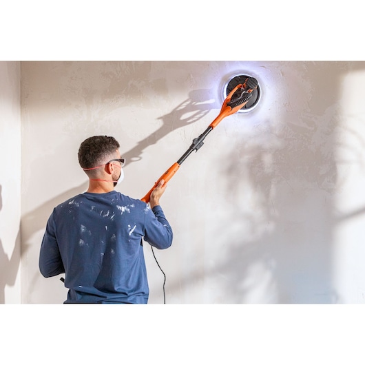 Person uses BLACK+DECKER BEDW700 electric wall sander with LED on a white wall, wearing mask and glasses.