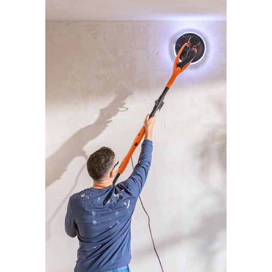Person using an orange BLACK+DECKER wall sander with LED light to sand a ceiling.
