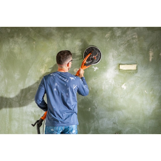 Person using a BLACK+DECKER wall sander to smooth a green painted wall during a renovation project.