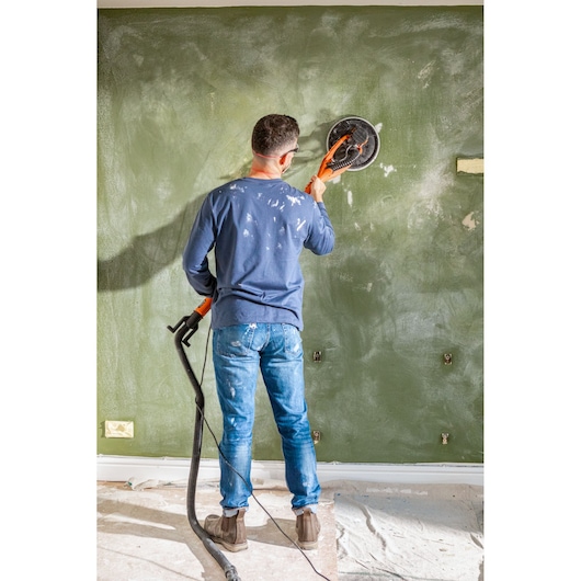 Person using a BLACK+DECKER wall sander on a green wall during renovation work.