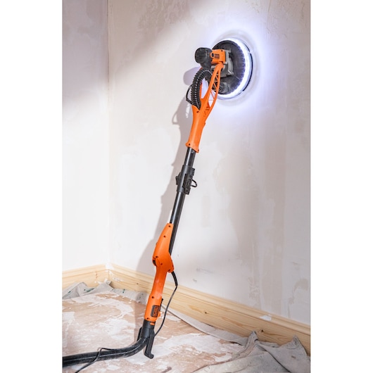 Orange BLACK+DECKER drywall sander with LED light sanding wall in a corner, cord and hose attached.