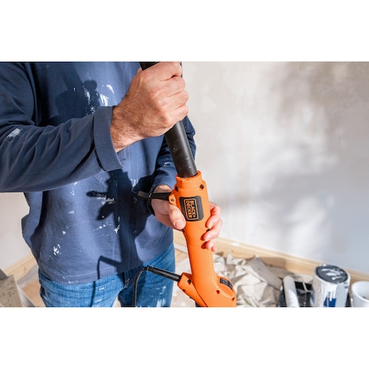 Person holding a BLACK+DECKER orange power tool in a room under renovation with paint cans on the floor.