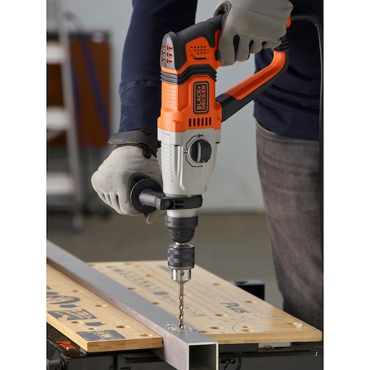 BLACKDECKER BEHS02K hammer drill used to drill metal on a workbench, operator wearing gloves.