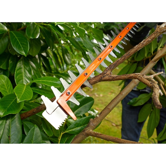 BLACKDECKER BEHTS451 hedge trimmer blade close-up cutting a thick tree branch among green leaves.