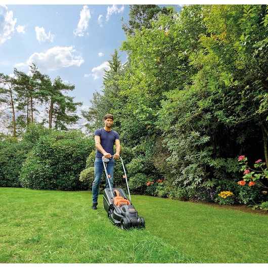 BLACKDECKER BEMW351GL2 lawn mower in use on green grass in a garden, with trees and flowers in background.