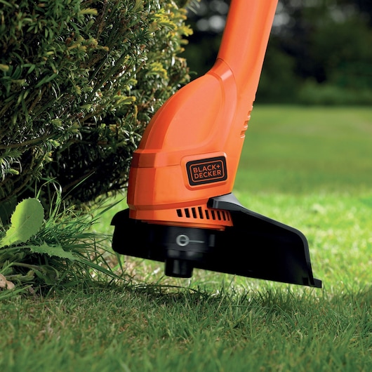 Photo of BLACK+DECKER BEMW461BGL2 orange grass trimmer in use trimming lawn edges near shrubs.