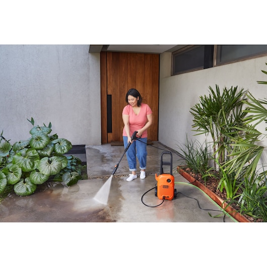 Person using BLACKDECKER BEPW1300L pressure washer to clean outdoor patio near plants and a wooden door.