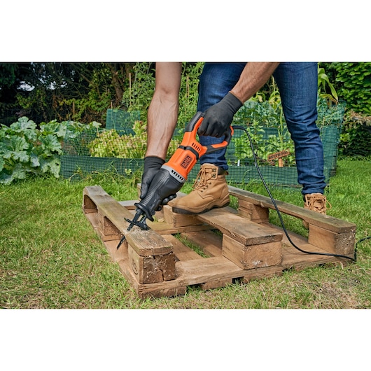 A person using a BLACK+DECKER reciprocating saw to cut through a wooden pallet outdoors. The person is wearing protective gloves and boots, and the saw is powered by a cord. There are plants and greenery in the background.