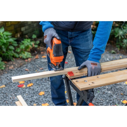 BLACKDECKER BES602 jigsaw in use, cutting wood on outdoor workbench, user wearing branded gloves.
