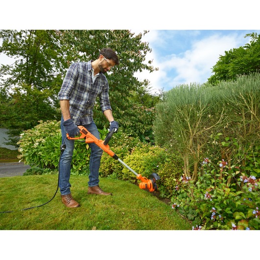 Photo of person using BLACKDECKER BESTA525 orange electric grass trimmer in a garden, trimming lawn edge.
