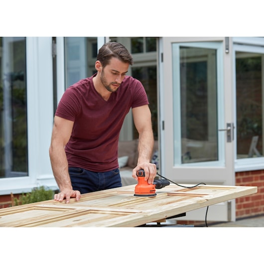 Person using BLACKDECKER BEW210 orange electric sander on a wooden door outdoors.