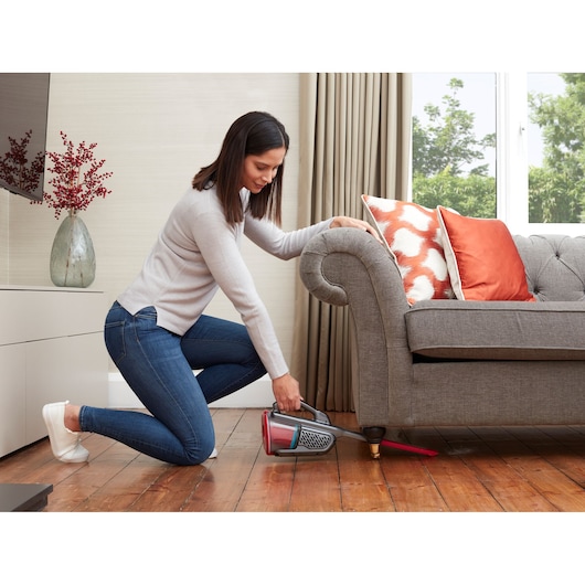 BLACKDECKER BHHV315J handheld vacuum cleaning under grey sofa with red accent pillows in living room.