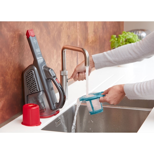 BLACKDECKER BHHV315J handheld vacuum filter being washed under a kitchen faucet, vacuum on counter.