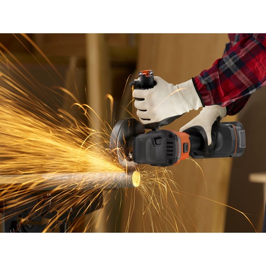 A person wearing protective gloves uses a BLACK+DECKER cordless angle grinder to cut through a metal pipe, creating bright, flying sparks.