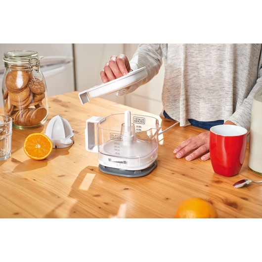 Person assembling a BLACK+DECKER citrus juicer on a kitchen table with oranges, cookies, glass and a red mug.