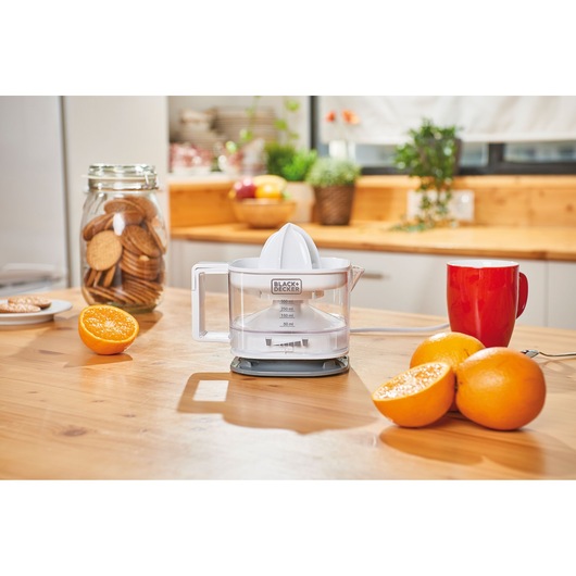 BLACK+DECKER BXCJ25E electric citrus juicer on wooden kitchen table with oranges, mug, and jar of cookies.