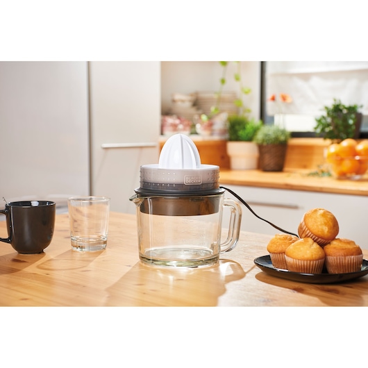 BLACK+DECKER BXCJ30E electric citrus juicer with glass jug on kitchen table near cup, glass, and plate of muffins.