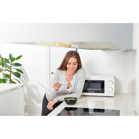 BLACK+DECKER BXMY700E white microwave oven on kitchen counter with a woman eating soup in front.
